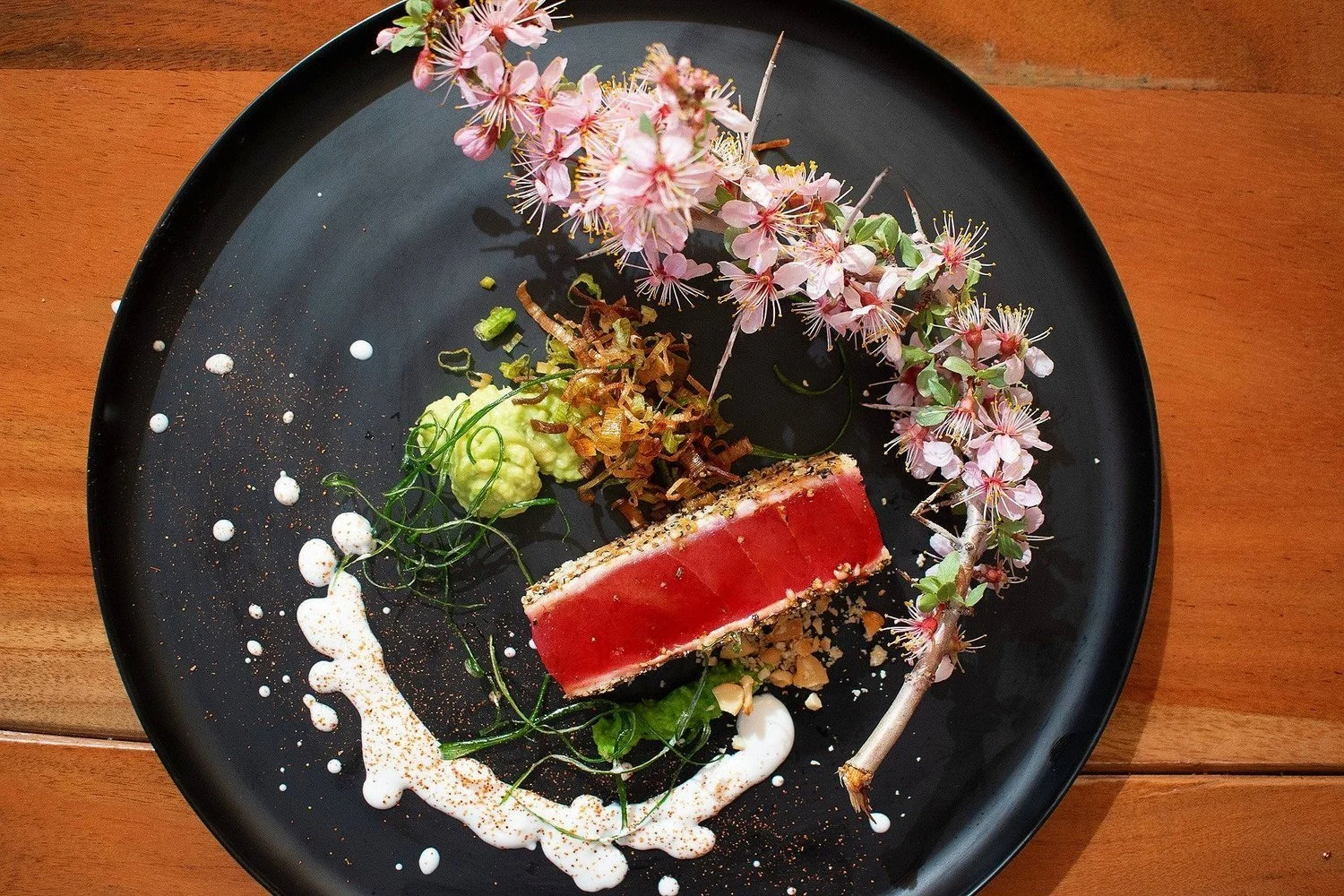 Artfully plated dish featuring a glazed protein, garnished with microgreens, cream or foam, crispy elements, and pink flowering sprigs on a black plate.