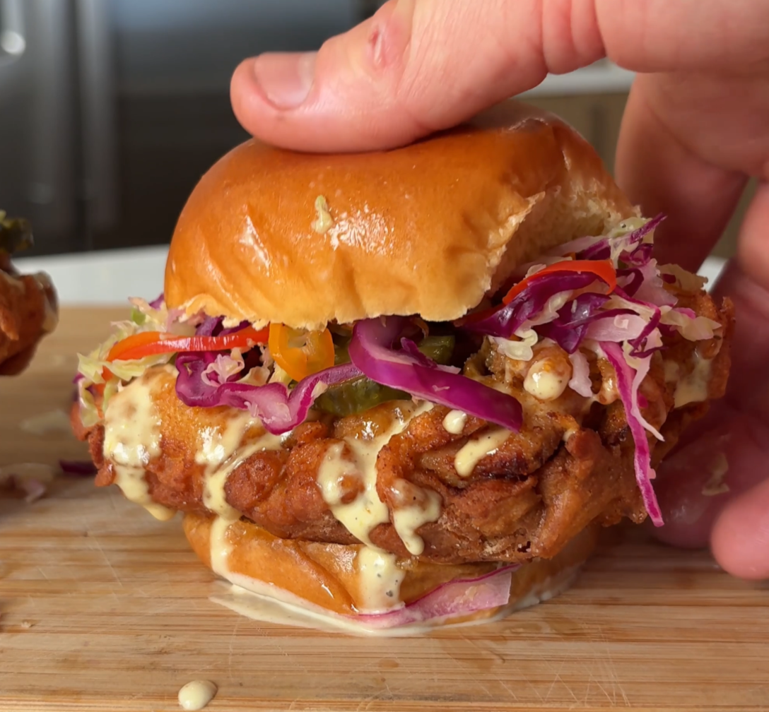 Hand holding a fried chicken sandwich with coleslaw, mustard, and a bun on a wooden cutting board.