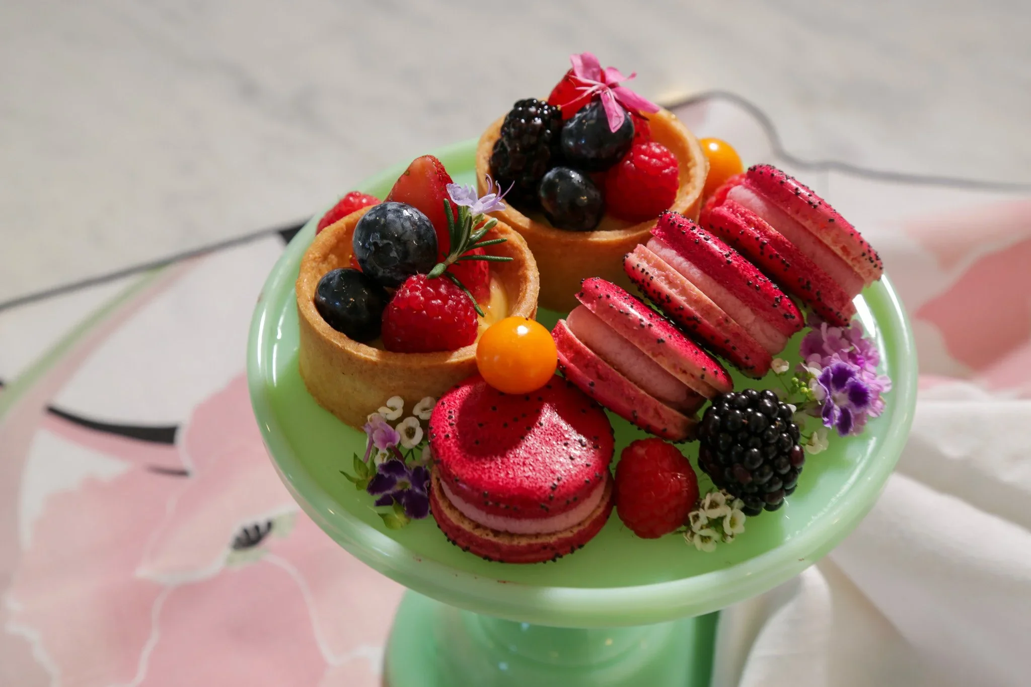A small green pedestal dish holding two fruit tarts filled with cream, topped with mixed berries and flowers, along with pink macarons decorated with black poppy seeds, also garnished with berries and flowers.