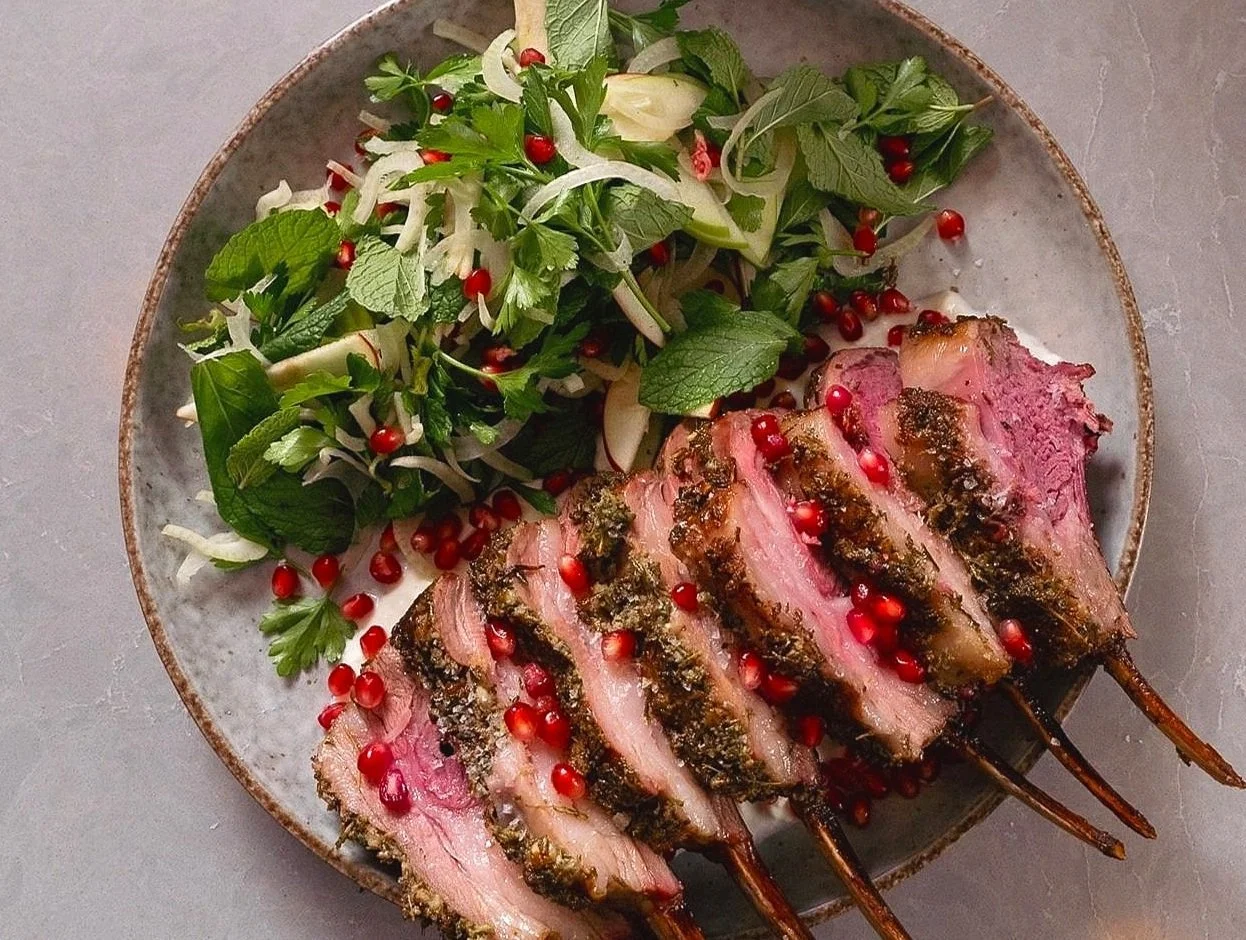 Plate of grilled lamb chops with herb crust, served with a side salad of mixed greens, herbs, sliced fennel, and pomegranate seeds.