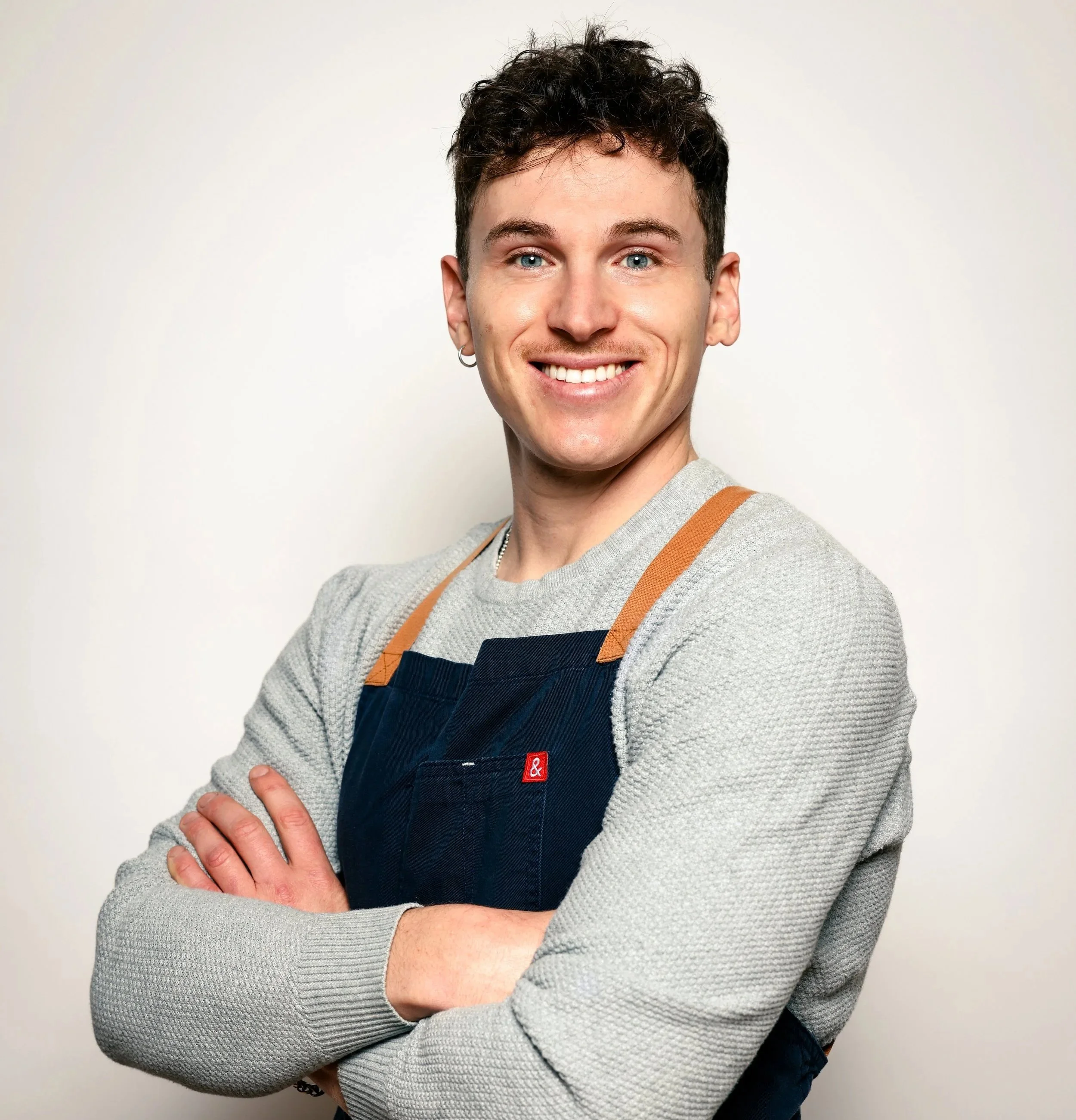 A young man with short curly dark hair, blue eyes, and light skin, smiling and standing with arms crossed, wearing a gray sweater and a dark apron with tan straps in front of a plain white background.
