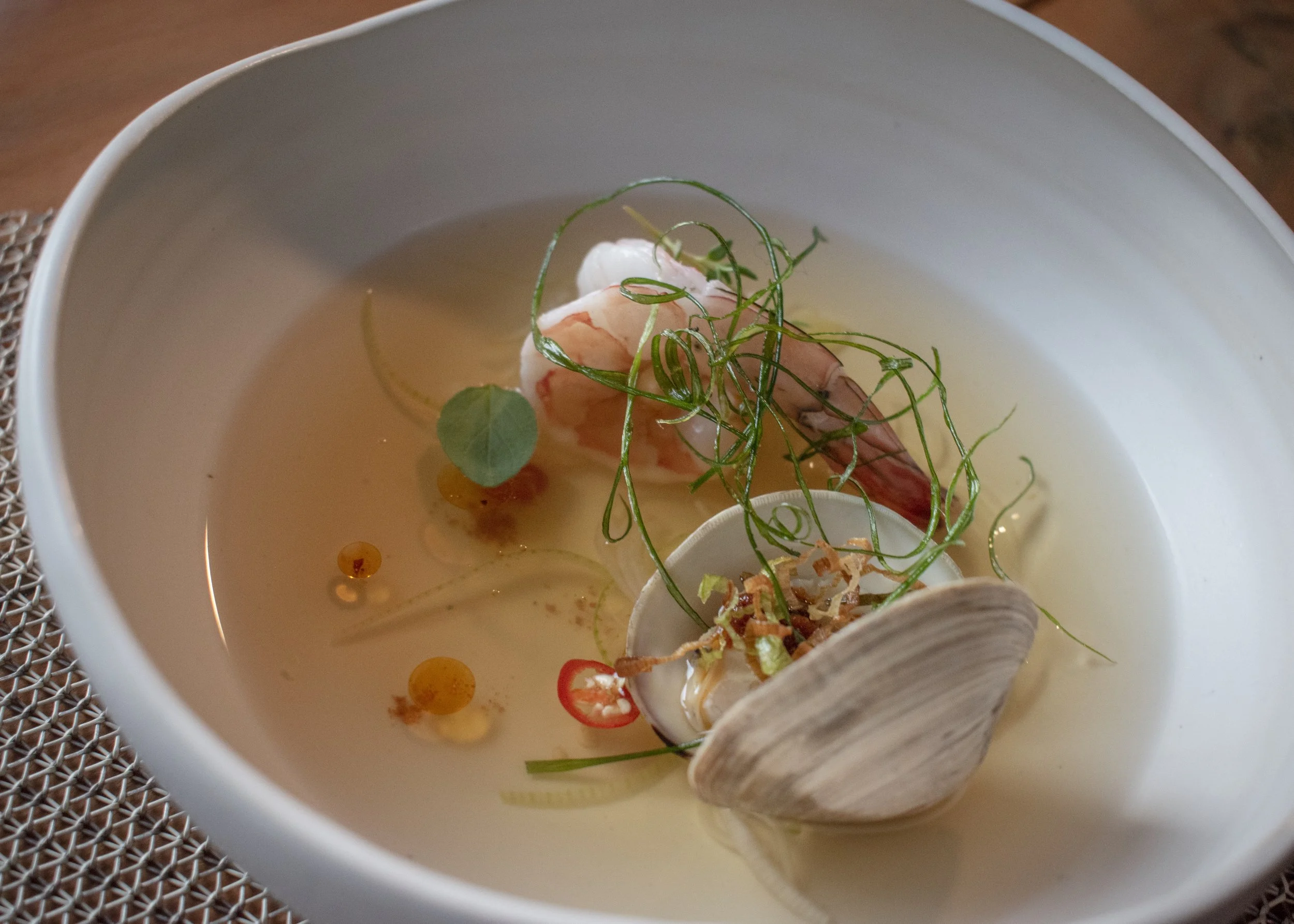 A bowl of seafood soup with a shrimp, garnished with herbs, a shell, and a slice of chili.