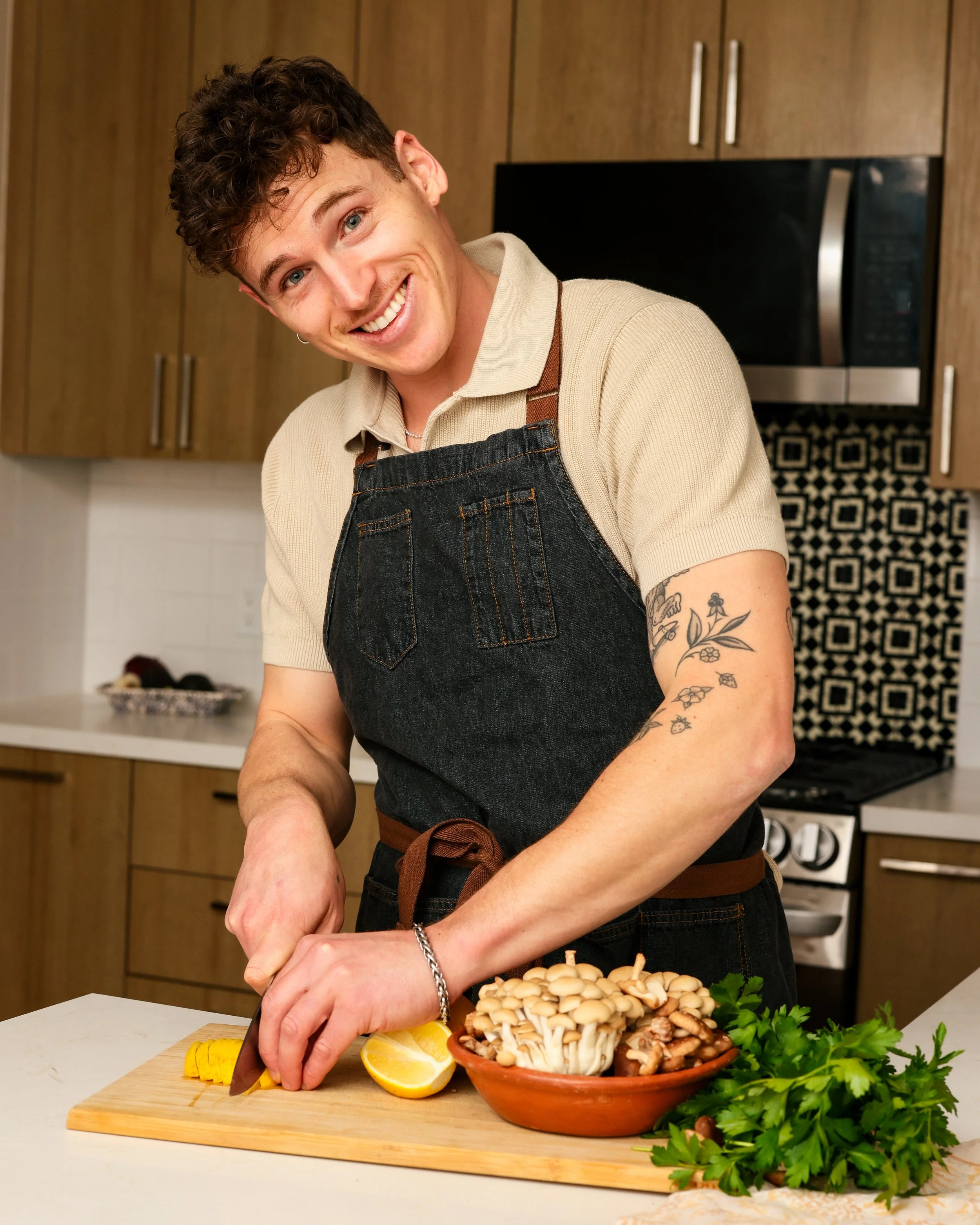 Smiling man preparing mushrooms and lemons in a modern kitchen with wooden cabinets, wearing a beige polo shirt, black apron, and a chain bracelet.