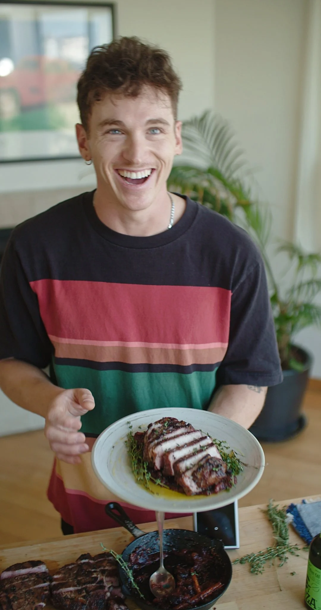 A young man with brown hair and blue eyes, smiling and holding a plate of sliced meat with a green sauce, standing in a bright room with a plant in the background.