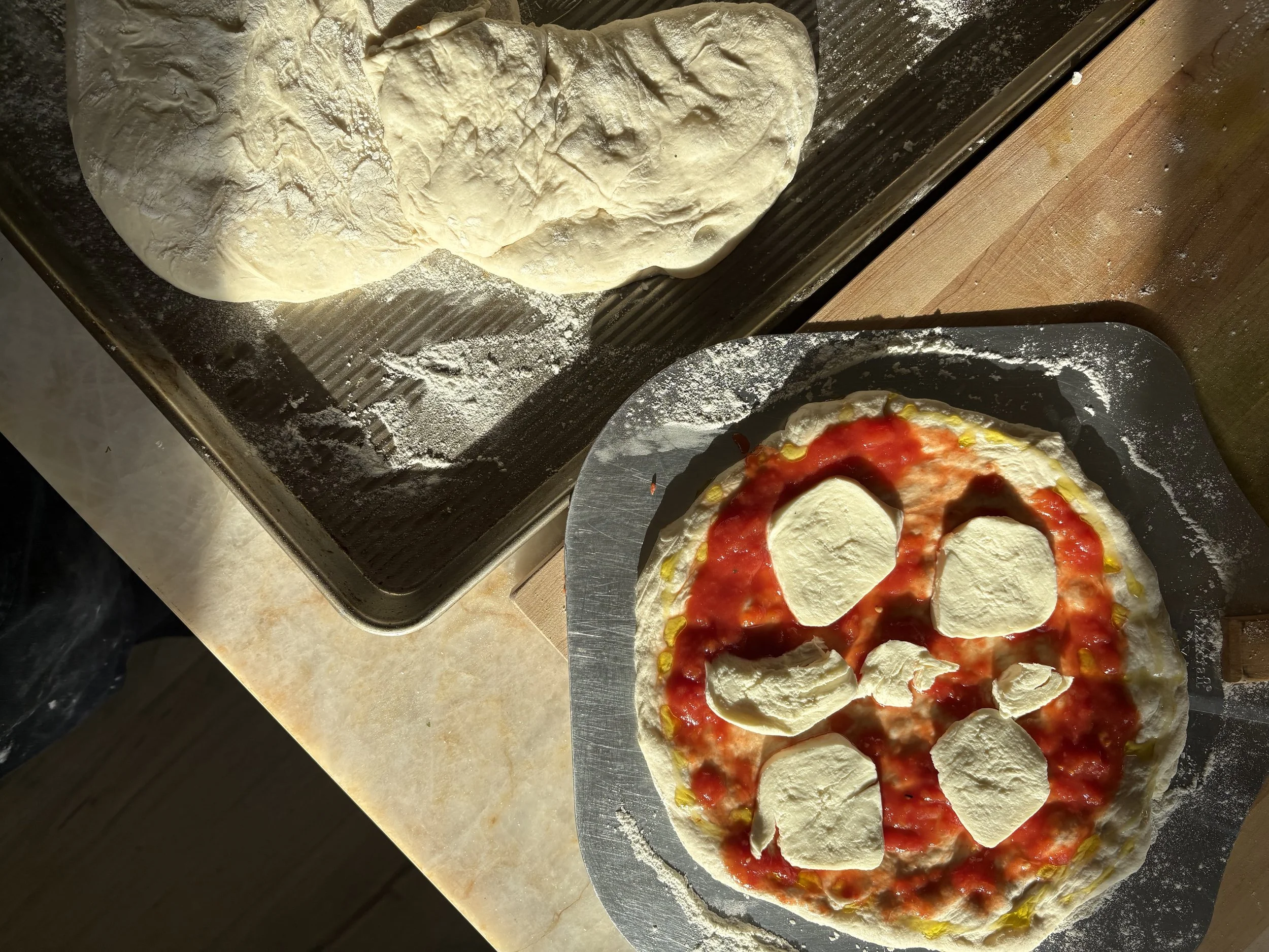 A pizza decorated with cheese slices, tomato sauce, and yellow bell peppers before baking, on a pizza peel, with a dough ball on a floured baking sheet.