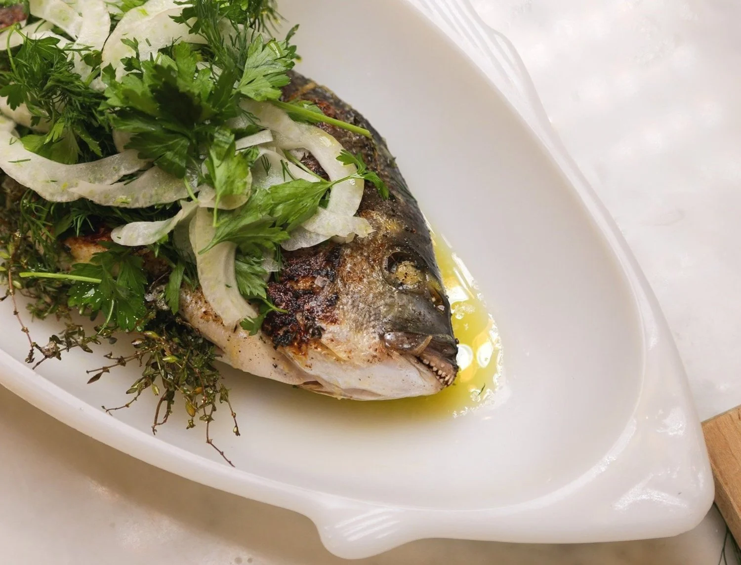 A cooked fish garnished with fresh herbs and sliced onions on a white platter with olive oil.