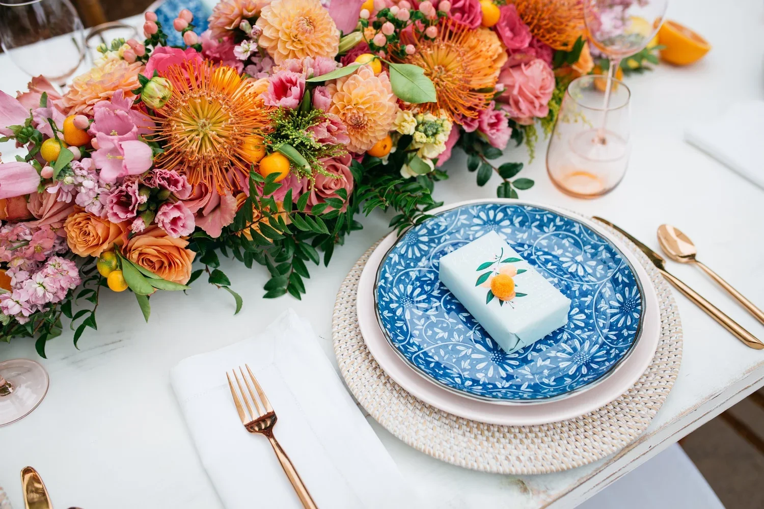 A table setting for a party with a large colorful flower arrangement centerpiece featuring pink, orange, and peach flowers with greenery. The table has a patterned blue plate with a small wrapped gift decorated with a peach motif on top, placed on a 