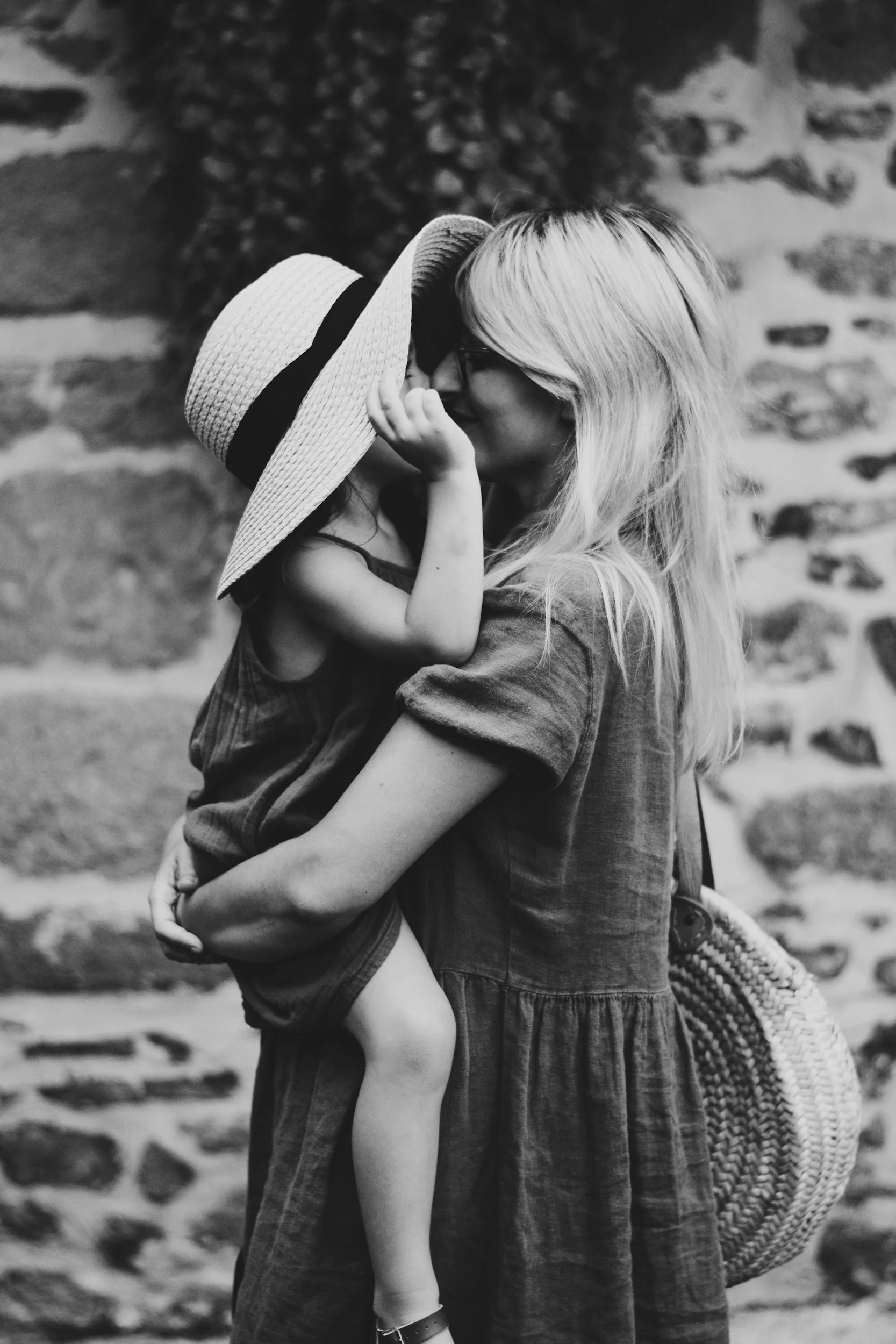 A woman with long blonde hair holding a young girl wearing a wide-brimmed hat, both leaning in close with their faces touching, in front of a stone wall.