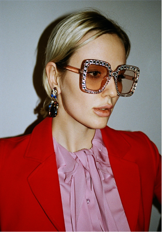 A woman with short blonde hair wearing large, square, rhinestone-encrusted sunglasses, a pink blouse with a bow at the collar, a red blazer, and large statement earrings.