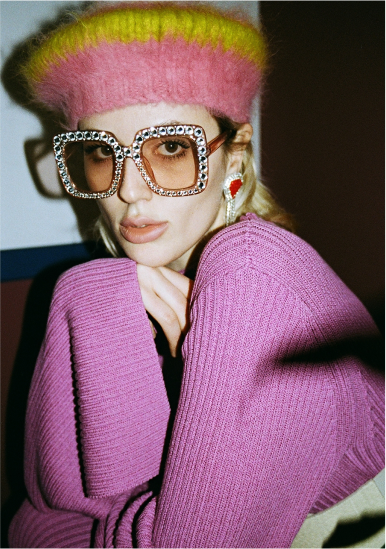 A woman wearing a multicolored fuzzy beret, large rhinestone-encrusted glasses, pink sweater, and statement earrings, posing with her hand near her chin.