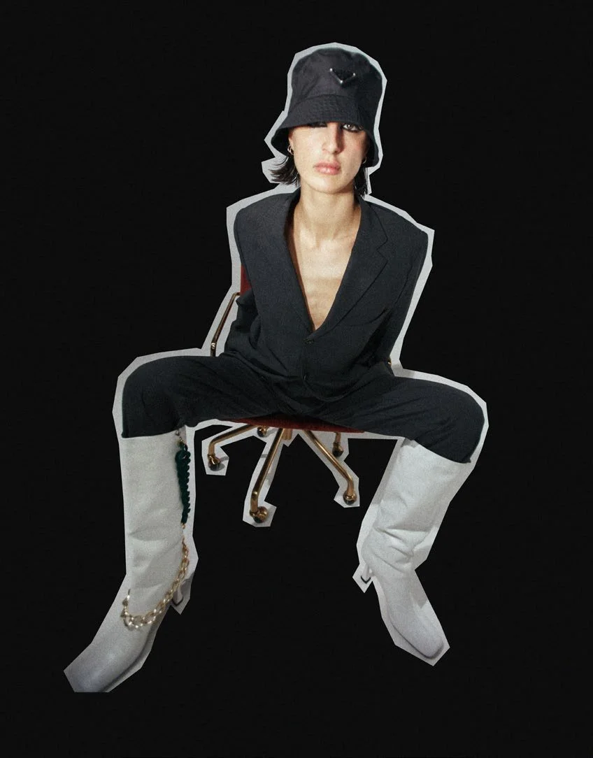 A woman sitting on a chair against a black background, wearing a black blazer, white knee-high boots with chains, and a black bucket hat.