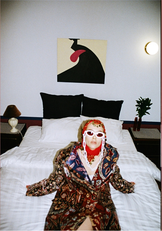 Person lying on a bed, dressed in colorful, patterned clothing with large sunglasses and jewelry, in a modern bedroom with abstract art on the wall and minimal decor.