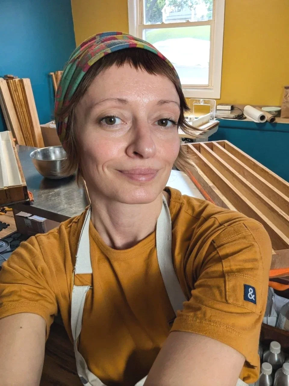 A woman taking a selfie in a woodworking workshop. She has short hair, is wearing a colorful headscarf, a mustard yellow shirt, and an apron. Behind her, there are wooden boxes or frames, a metal bowl, and various woodworking supplies on the counter.