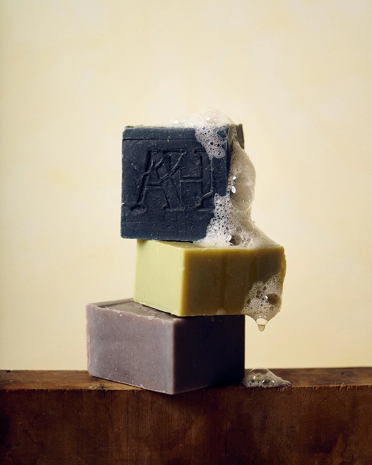 Stacked bars of soap in gray, yellow, and dark blue colors on a wooden surface with soap bubbles on top.