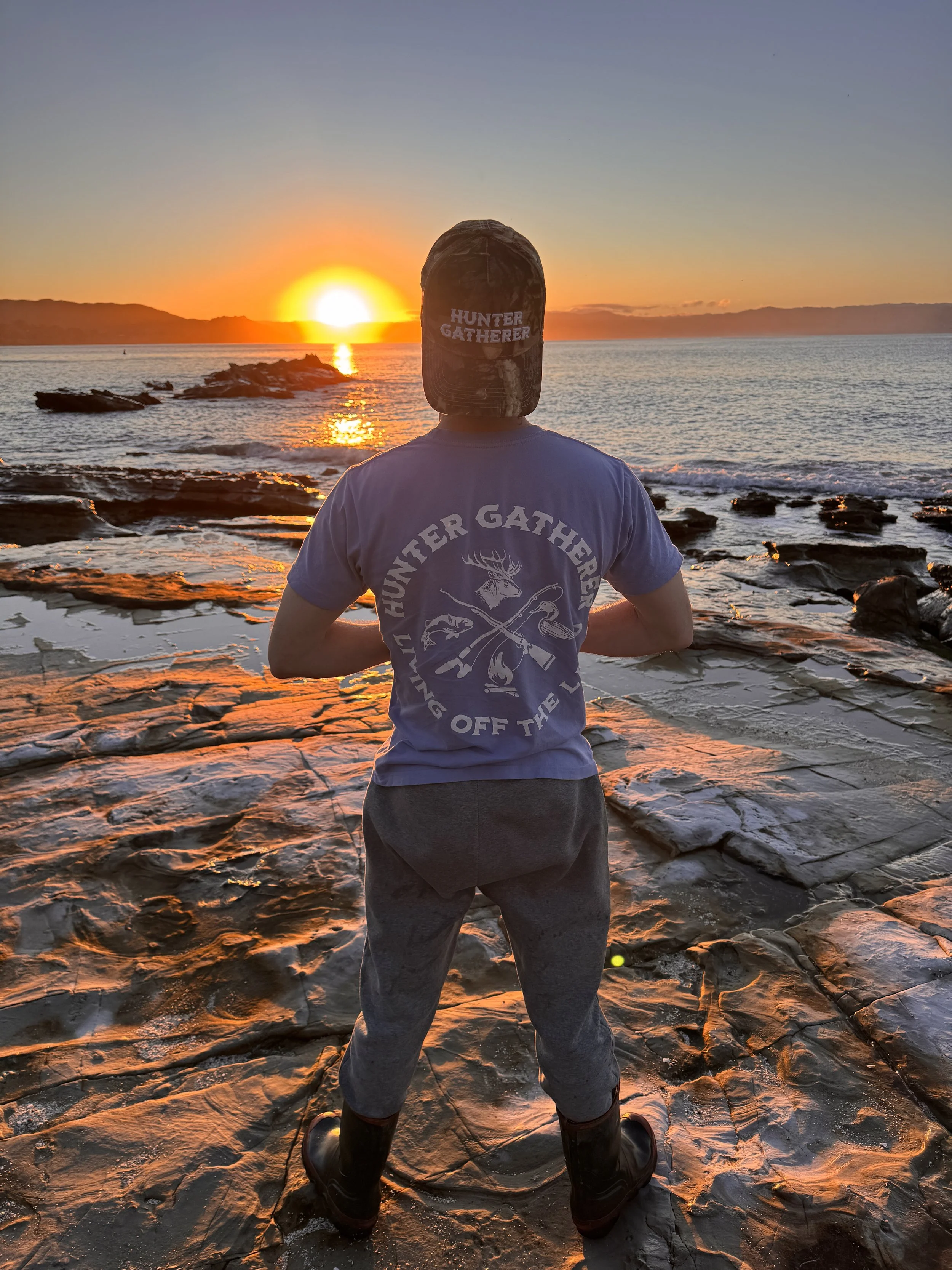 A person wearing a cap and t-shirt, standing on wet rocks at the beach during sunset, facing the sunset over the water.