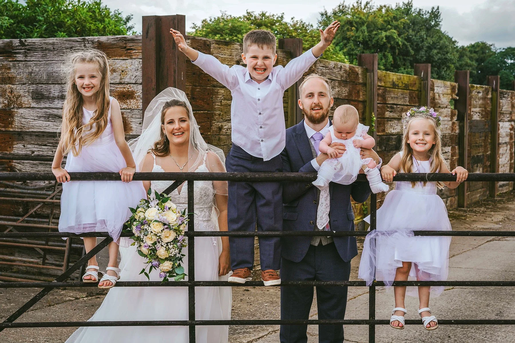 smiling children stand on cattle gate smiling with bride and groom at manor farm barn wedding venue taken by david lefebvre