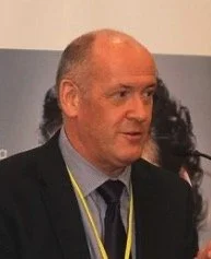 Man with a balding head and long hair at the sides, dressed in a dark suit and tie, at an indoor event or conference.