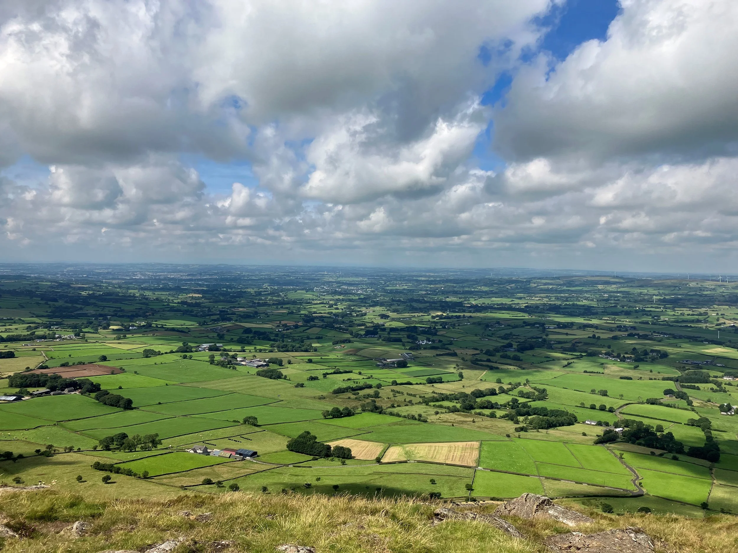 A vast green landscape with patchwork farmland, scattered trees, and a rural area under a partly cloudy sky.