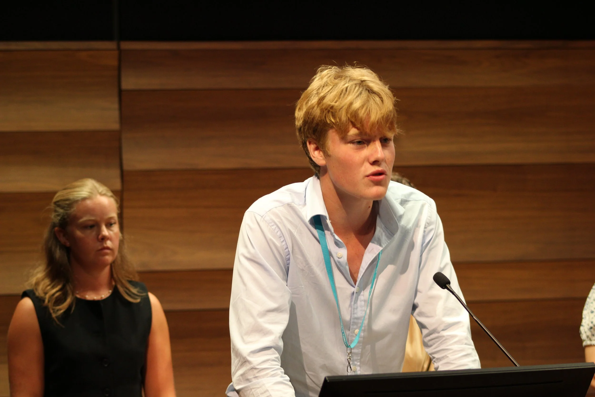 Young man with red hair, wearing a white shirt, speaking at a podium during a conference or meeting, with a microphone and a woman with long blonde hair and a black dress sitting beside him, against a wood-paneled wall.