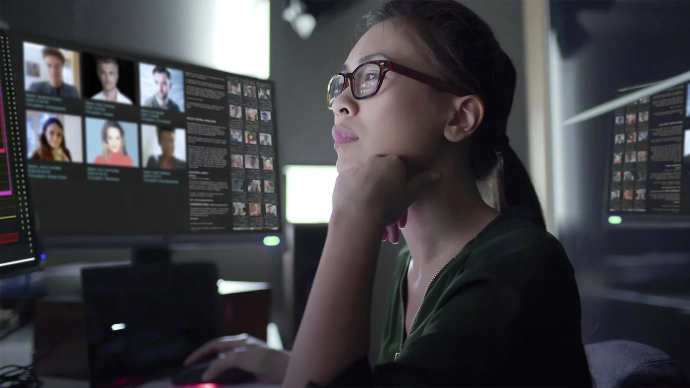 A woman with glasses sits at a desk, looking at multiple computer monitors displaying photos and text.