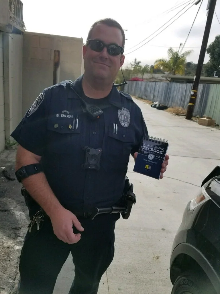 A police officer wearing sunglasses and a uniform, holding a small notebook that reads 'FacBook' and 'Field Resource,' standing outside next to a parked car in an alleyway.