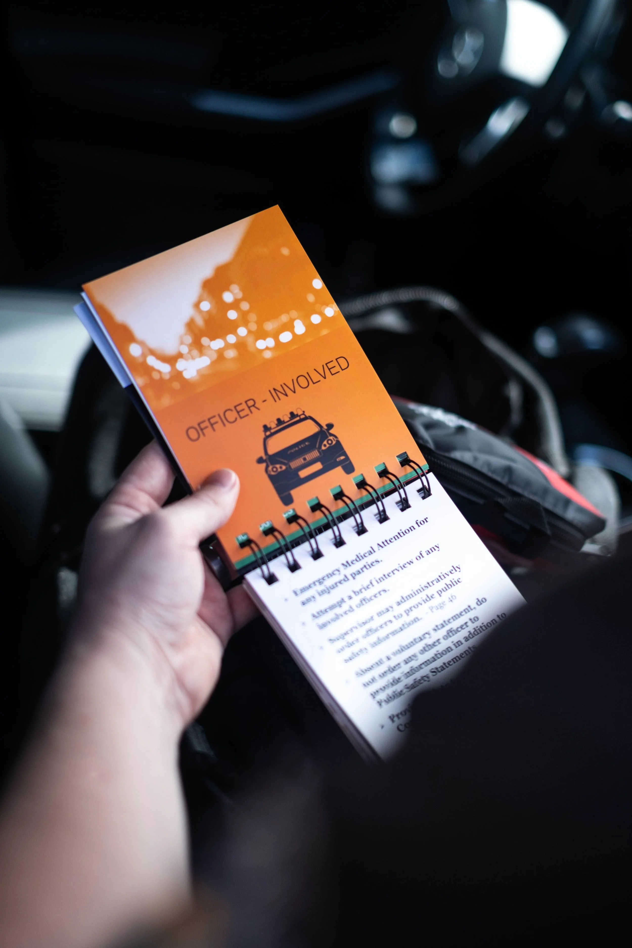 Hand holding a small booklet titled 'Officer - Involved' with a police car illustration on the cover inside a vehicle.