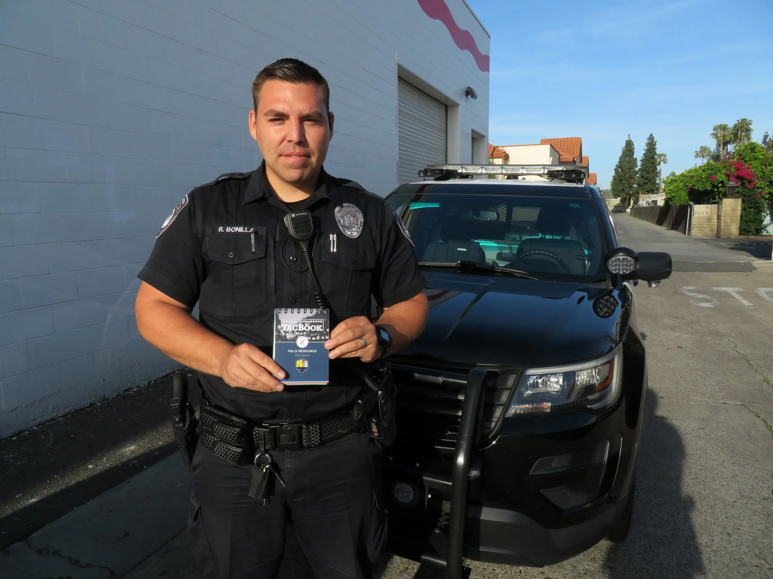 A police officer standing in front of a patrol car outdoors during the day, holding a Field Resource notebook.