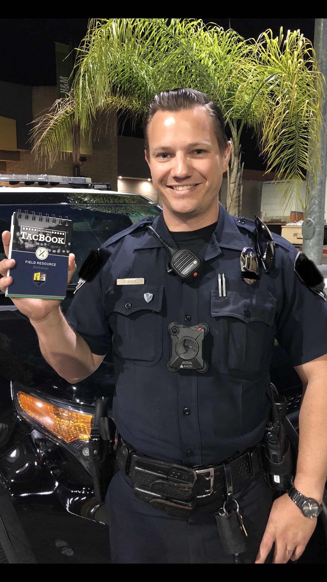 A smiling police officer in uniform holding a small notebook labeled 'TacBook' and standing in front of a black patrol vehicle with a palm tree in the background.