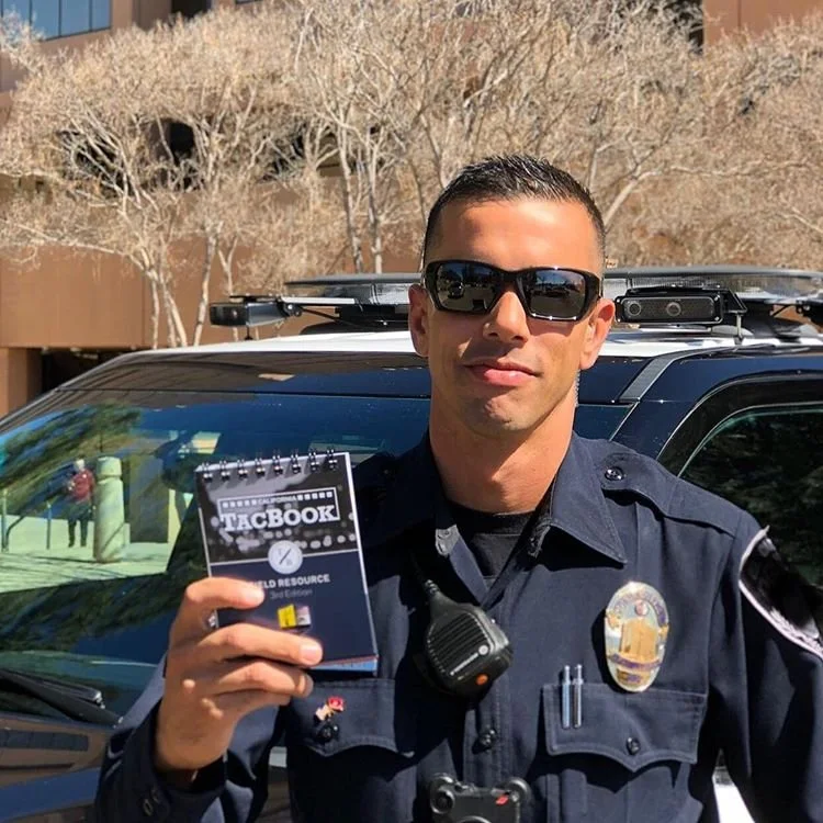A uniformed police officer wearing sunglasses holding a small notebook labeled 'TACBOOK' and standing next to a police vehicle outdoors with trees in the background.