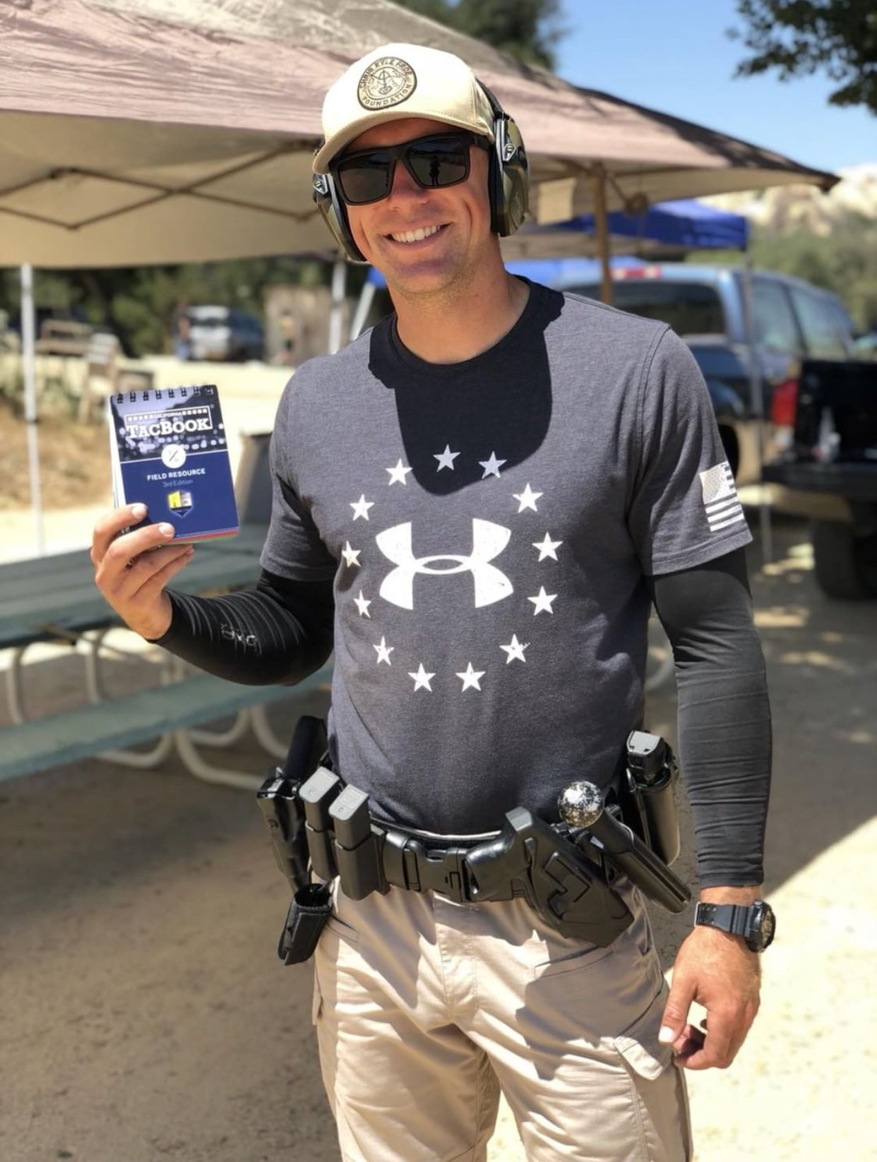 Man wearing sunglasses, a cap, and a tactical belt, holding a small notepad, smiling outdoors under a canopy with vehicles and trees in the background.