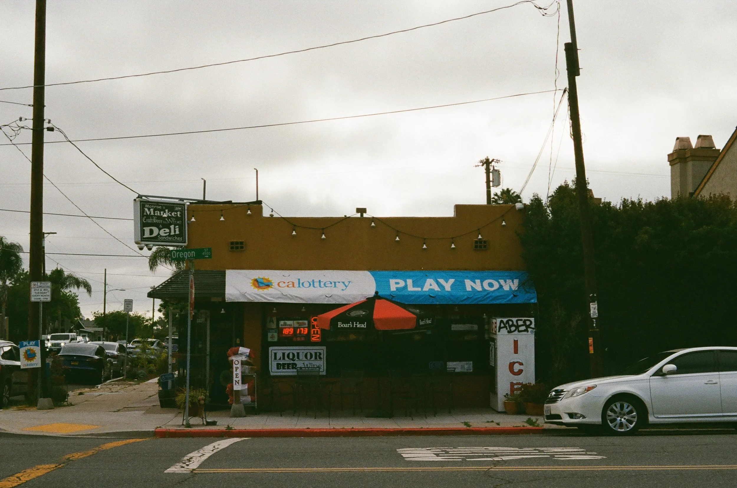 A small strip mall with a deli, a lottery kiosk, and a patio with an orange umbrella and string lights. Cars are parked in front, and utility poles and power lines are visible against a cloudy sky.