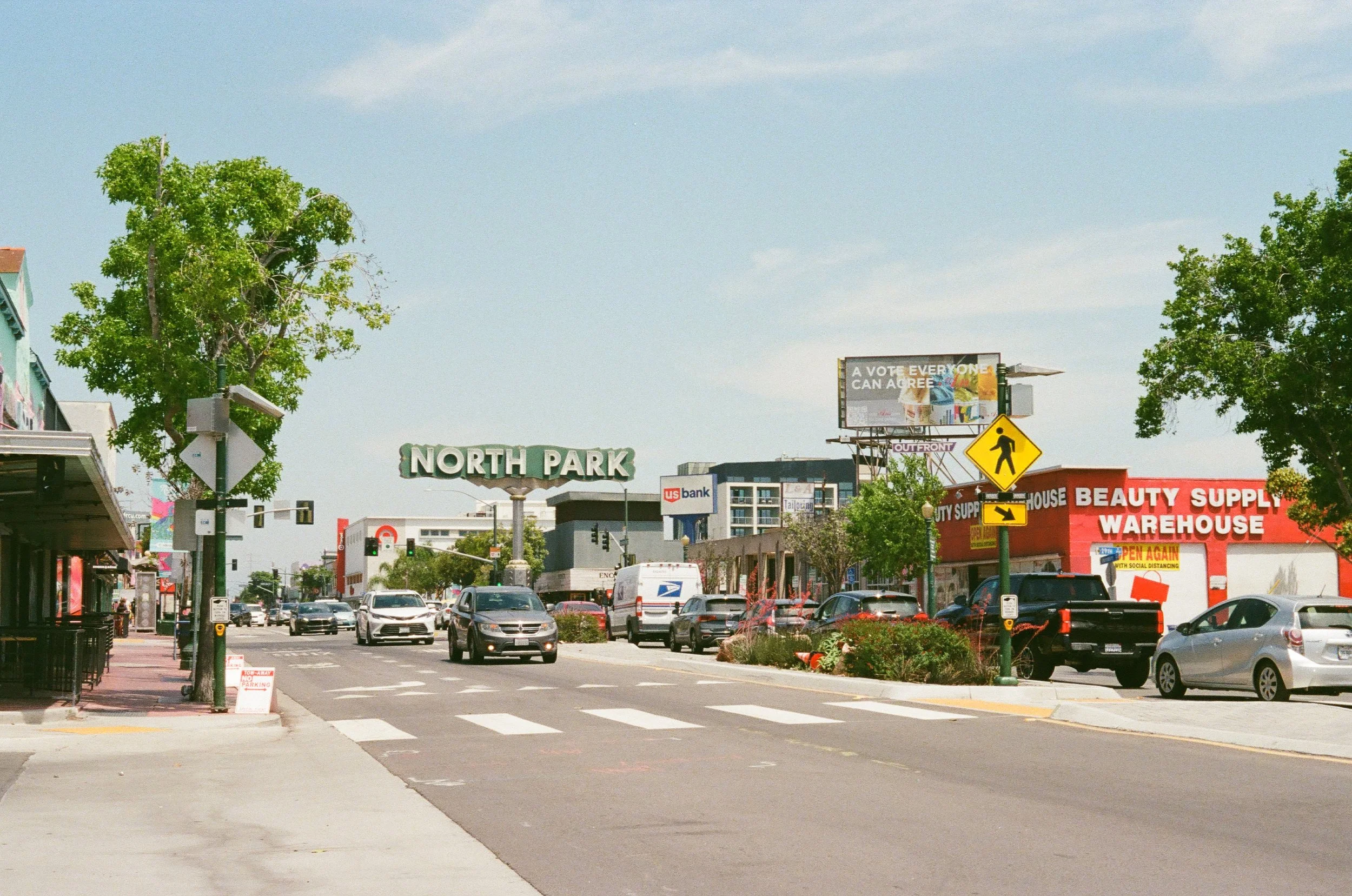 City street scene with cars, trees, storefronts, and overhead signs including a North Park sign, a USC bank sign, and a billboard that reads 'A vote everyone can agree.'