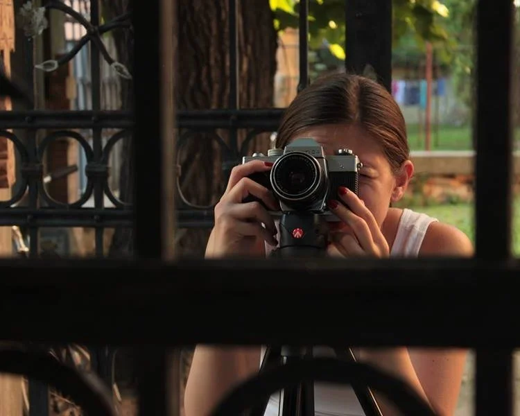 Girl taking a photo through a gate with a camera, with trees and greenery in the background.
