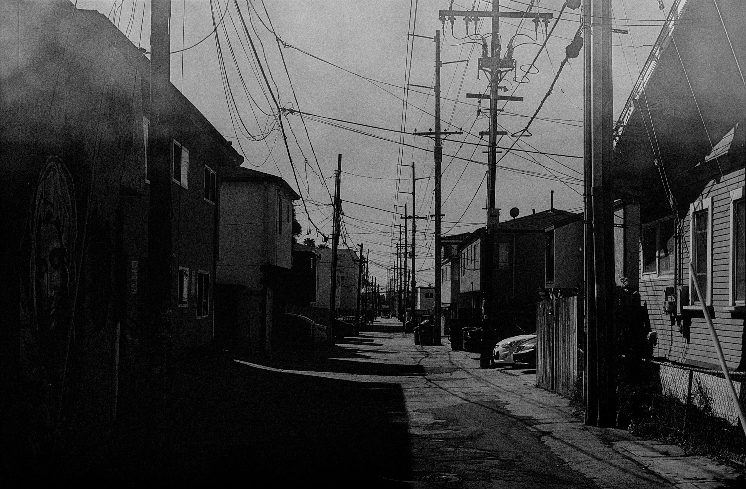 A black and white photo of a residential street with utility poles and a tangle of power lines, with houses on both sides and a car parked at the curb.