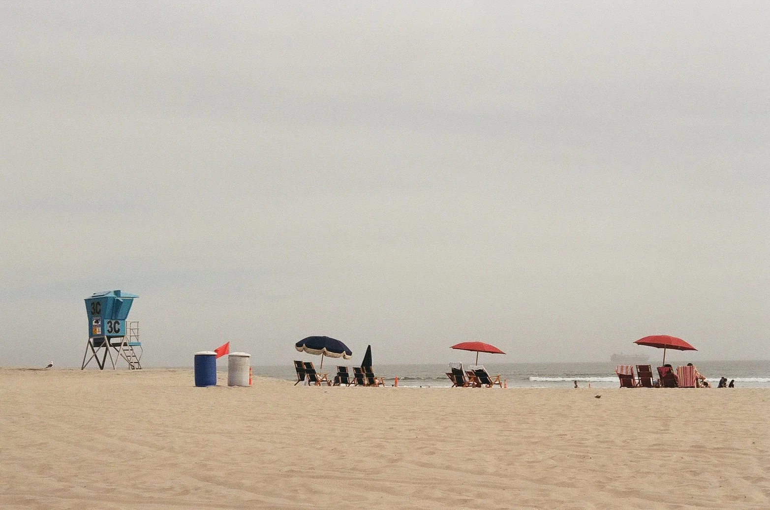 Empty beach with a lifeguard tower, chairs with umbrellas, and a few people near the shoreline on a cloudy day.