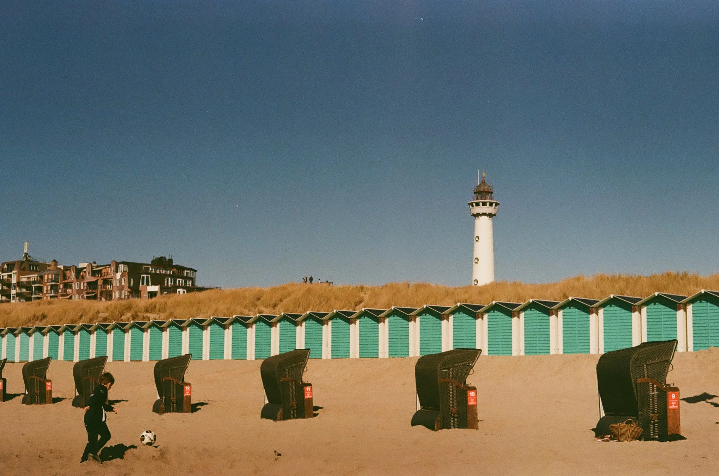 Children playing soccer on a sandy beach, with beach chairs and a row of beach huts in the foreground, a grassy hill with houses in the background, and a lighthouse on the hilltop under a clear blue sky.