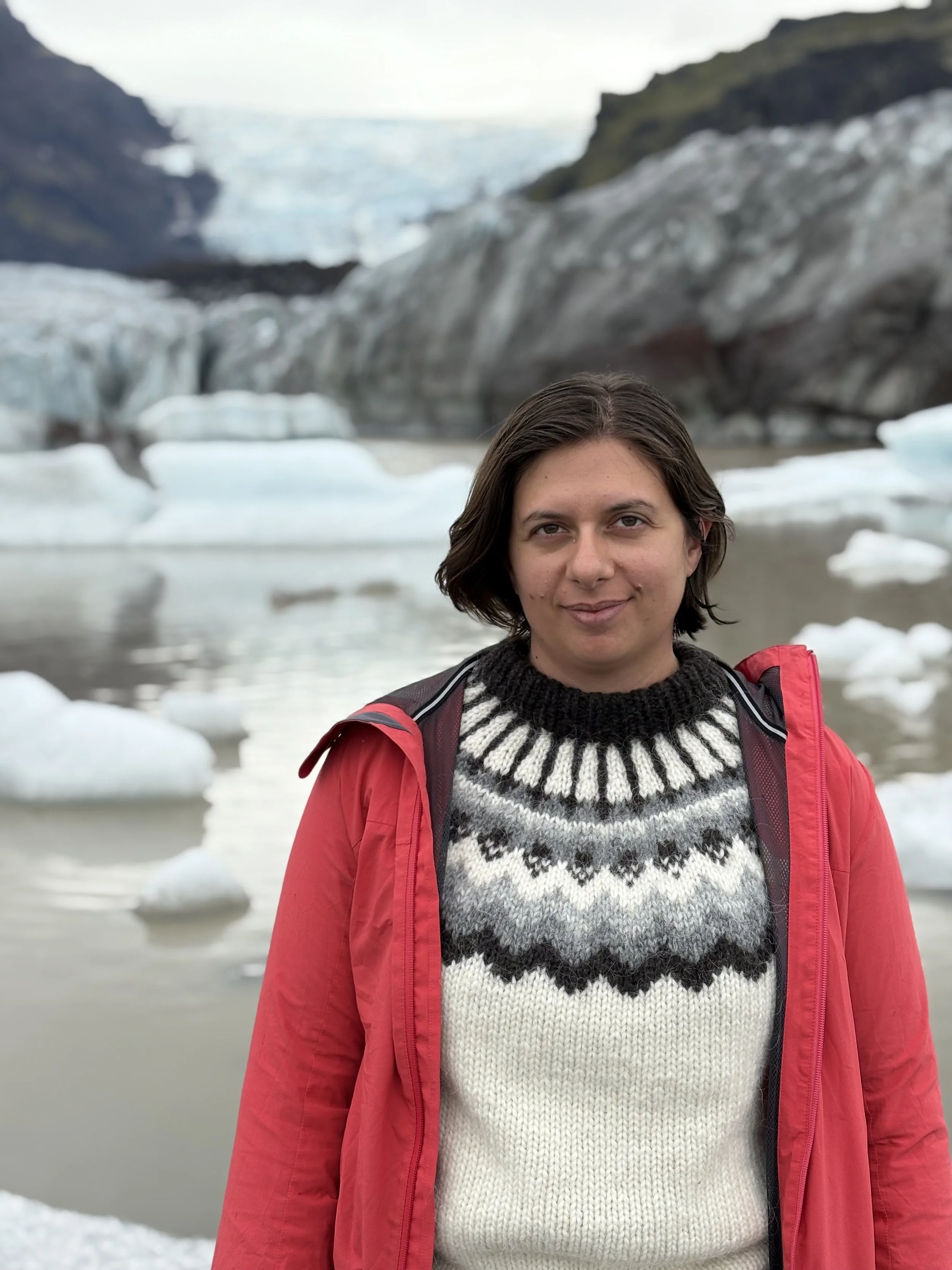 A young woman with short brown hair wearing a black and white patterned sweater and a red jacket stands in front of a glacier, mountain, and icy landscape with floating ice chunks in the water.
