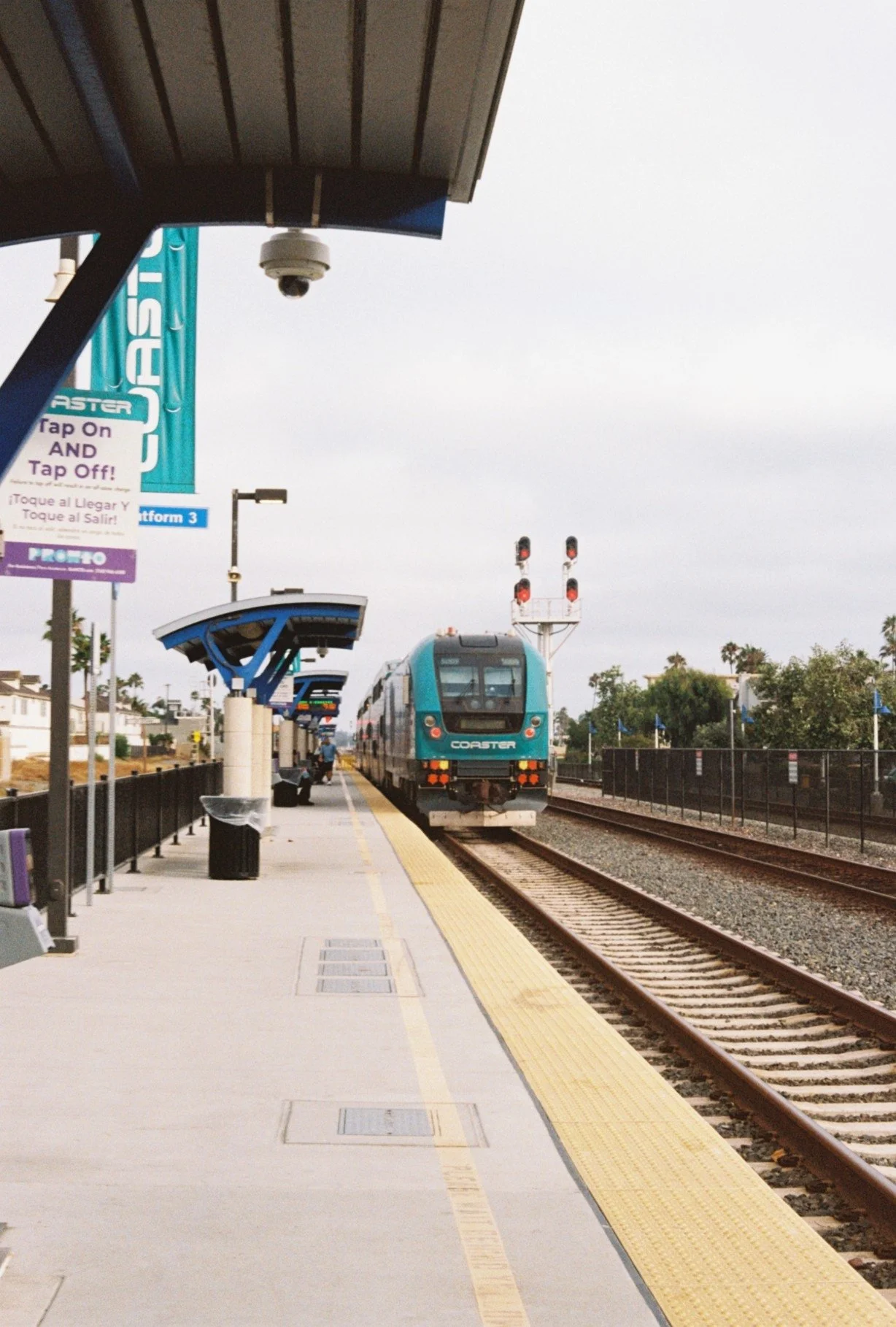 A train station platform with a blue modern train arriving on the tracks. The platform has a yellow tactile warning strip, benches, and signs, with some trees and buildings in the background. Overhead shelter provides shade.