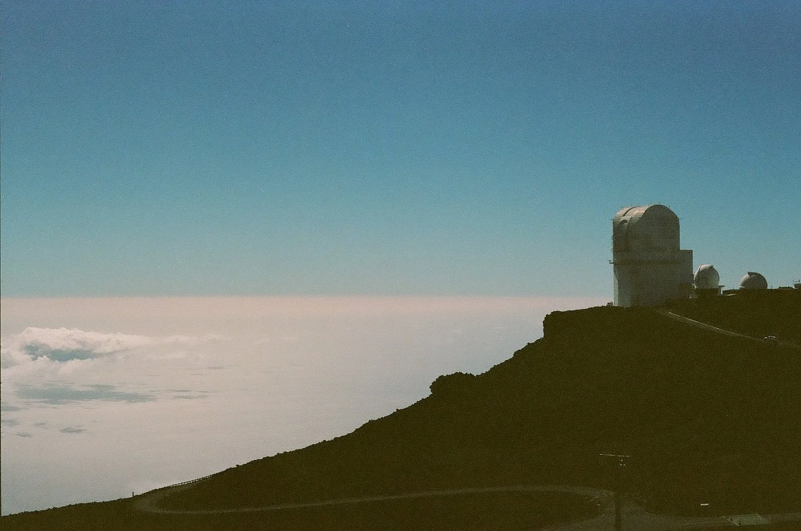 A mountain landscape with a clear blue sky, clouds below, and an observatory with several telescopes on the mountain peak.