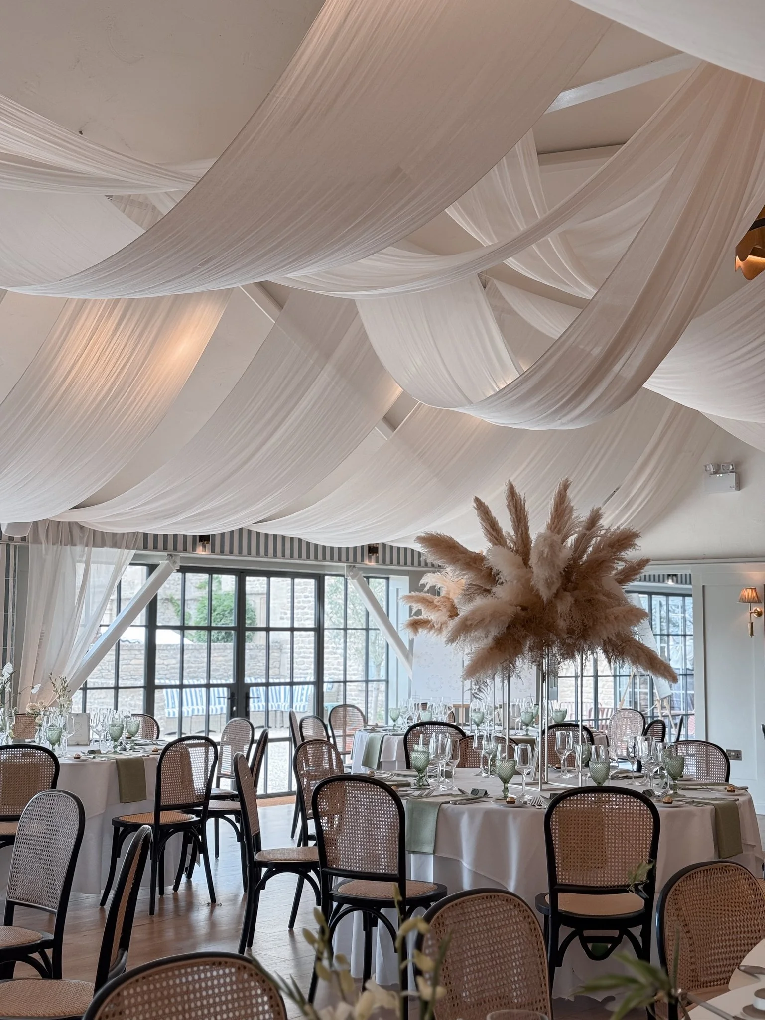 Elegant banquet or event room with draped white fabric on the ceiling, large windows with outdoor patio visible, round tables with white tablecloths, a centerpiece of pampas grass in a tall vase, and chairs with woven backs.