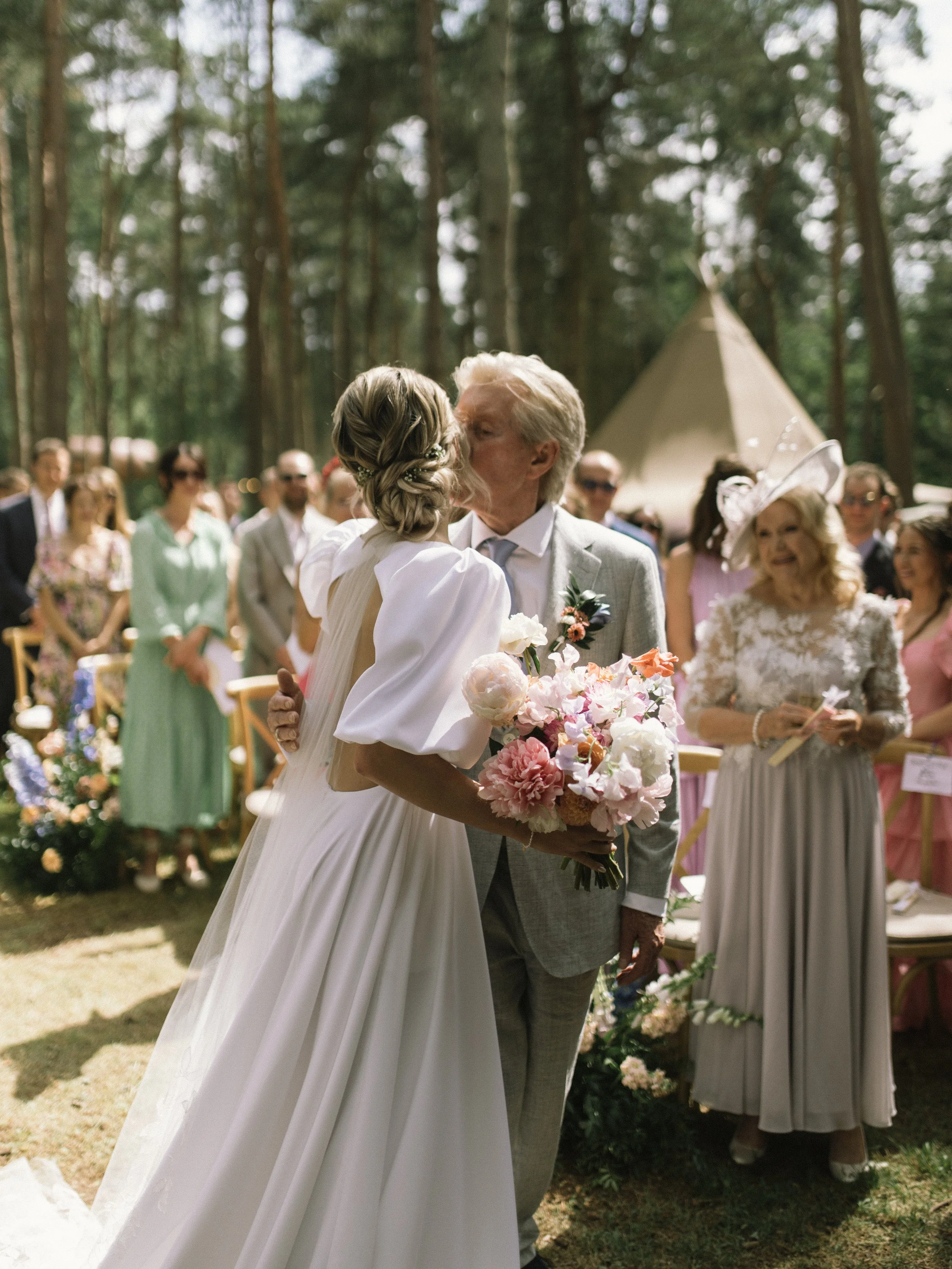 A bride and an elderly man sharing a kiss at an outdoor wedding ceremony in a wooded area, with guests in the background.