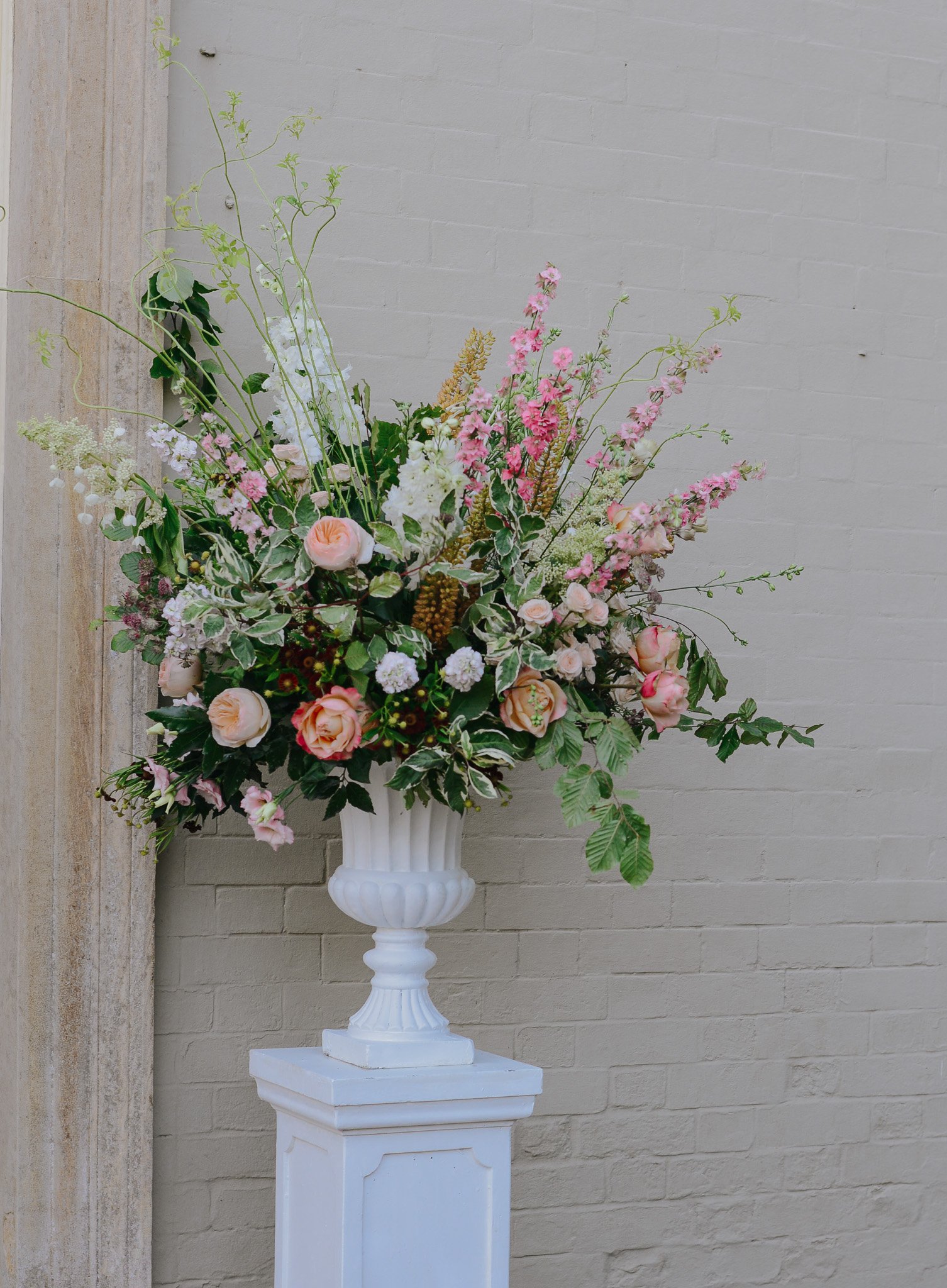 A large floral arrangement in a white vase on a white pedestal against a beige brick wall.