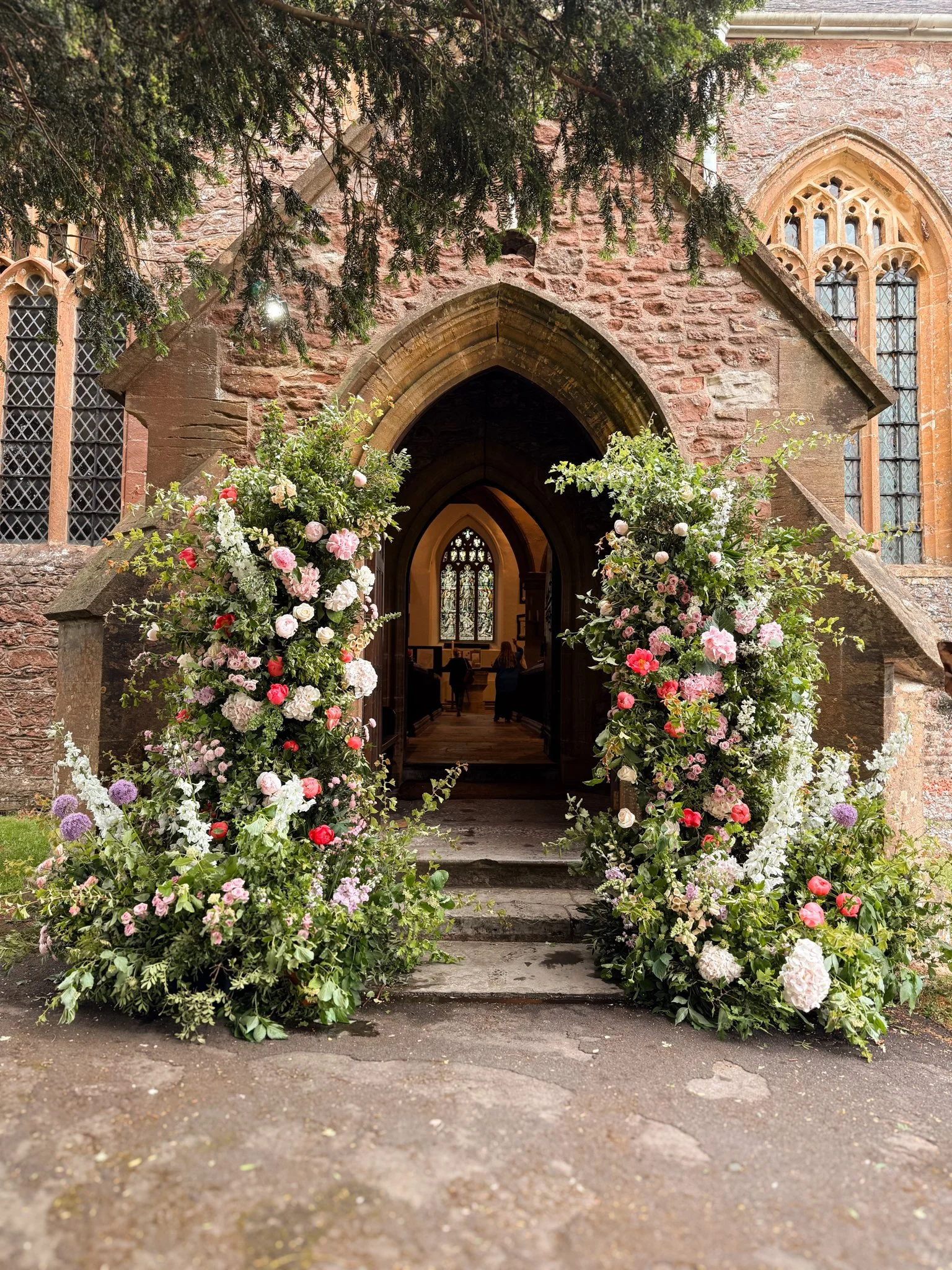 Floral archway with pink, white, and purple flowers at the entrance of a stone church with stained glass windows.