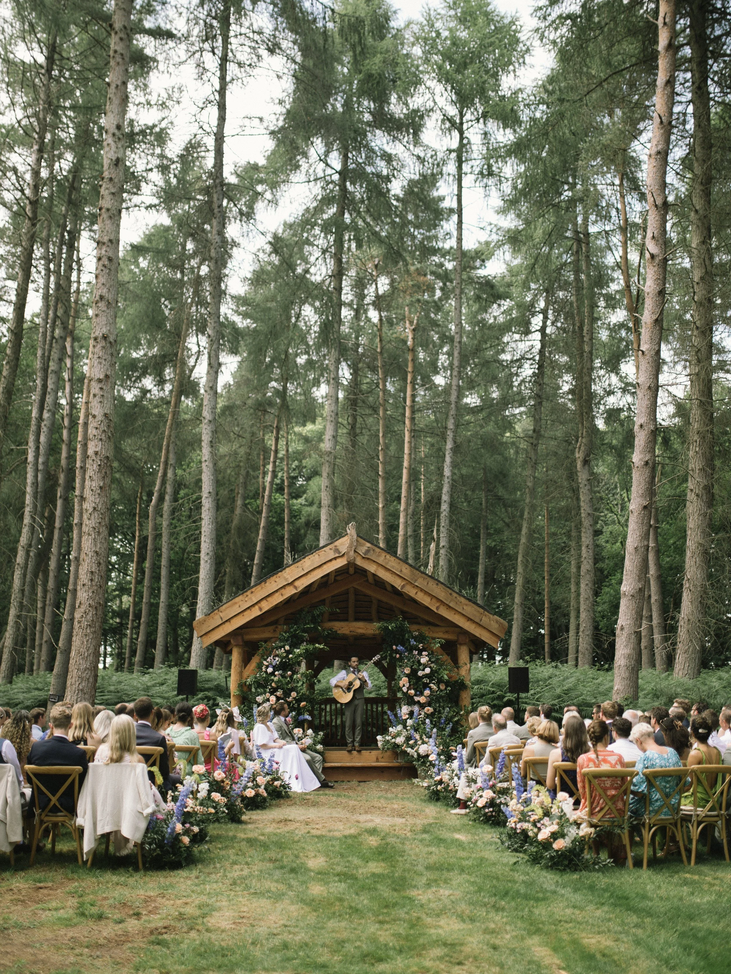 Outdoor wedding ceremony in a forest with a small wooden pavilion decorated with flowers, where a musician is playing guitar for seated guests.