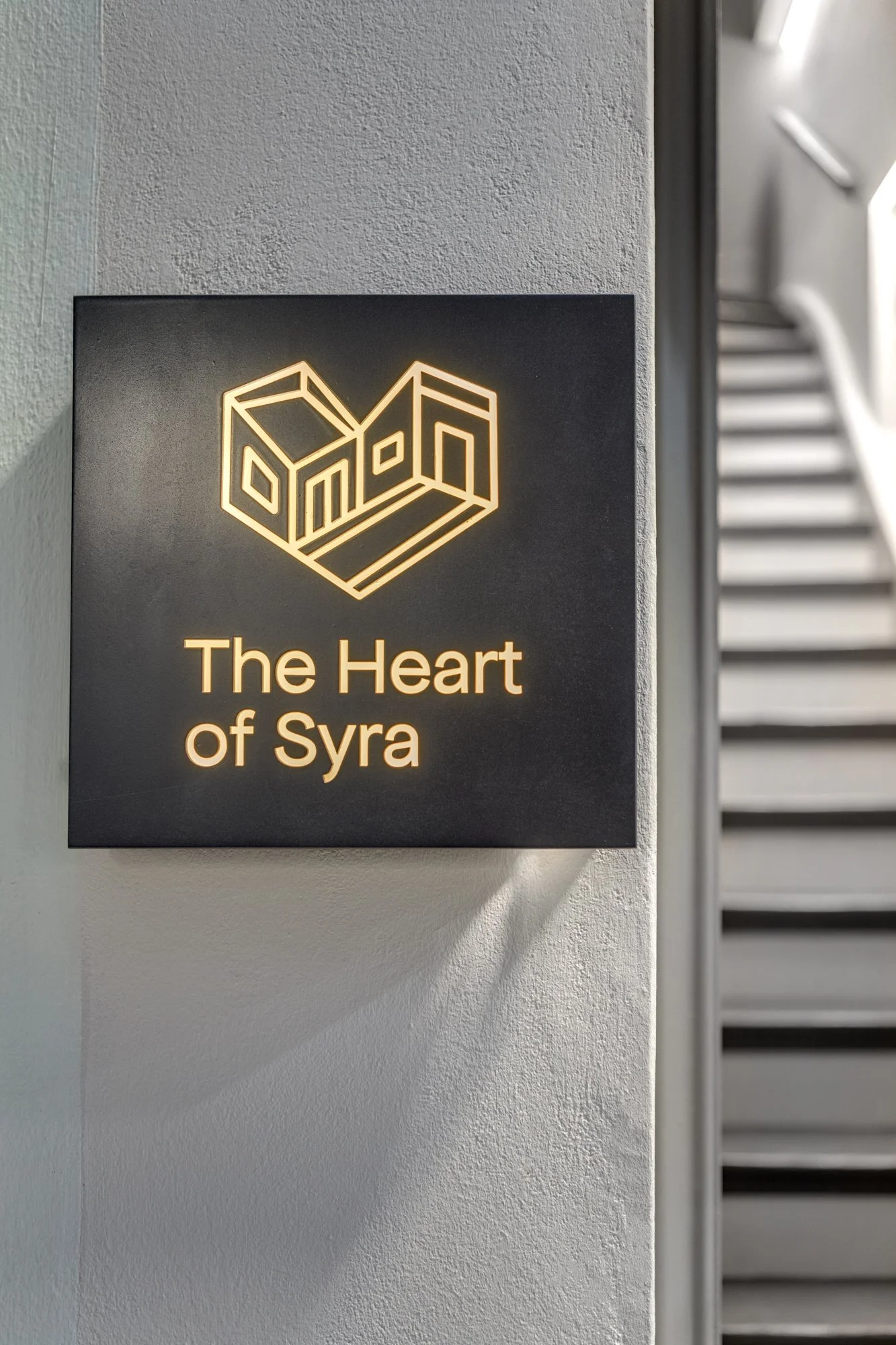Sign with a geometric heart logo and the text "The Heart of Syra" illuminated in warm light, mounted on a wall near a staircase.