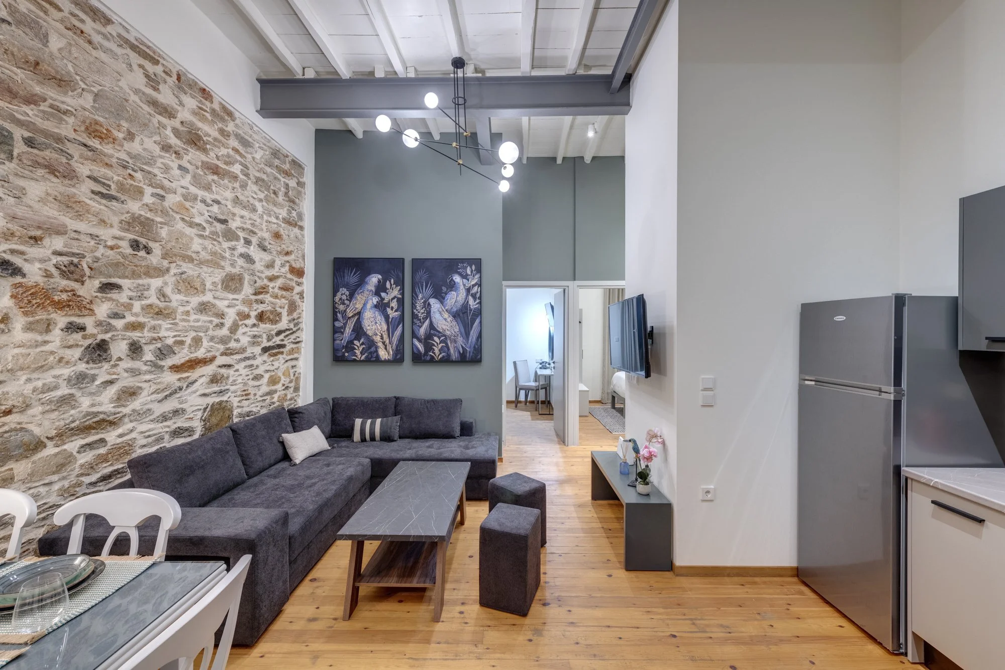 Living room with exposed brick wall, gray sectional sofa, wall art featuring parrots, flat-screen TV, modern chandelier, and dining area in the background.