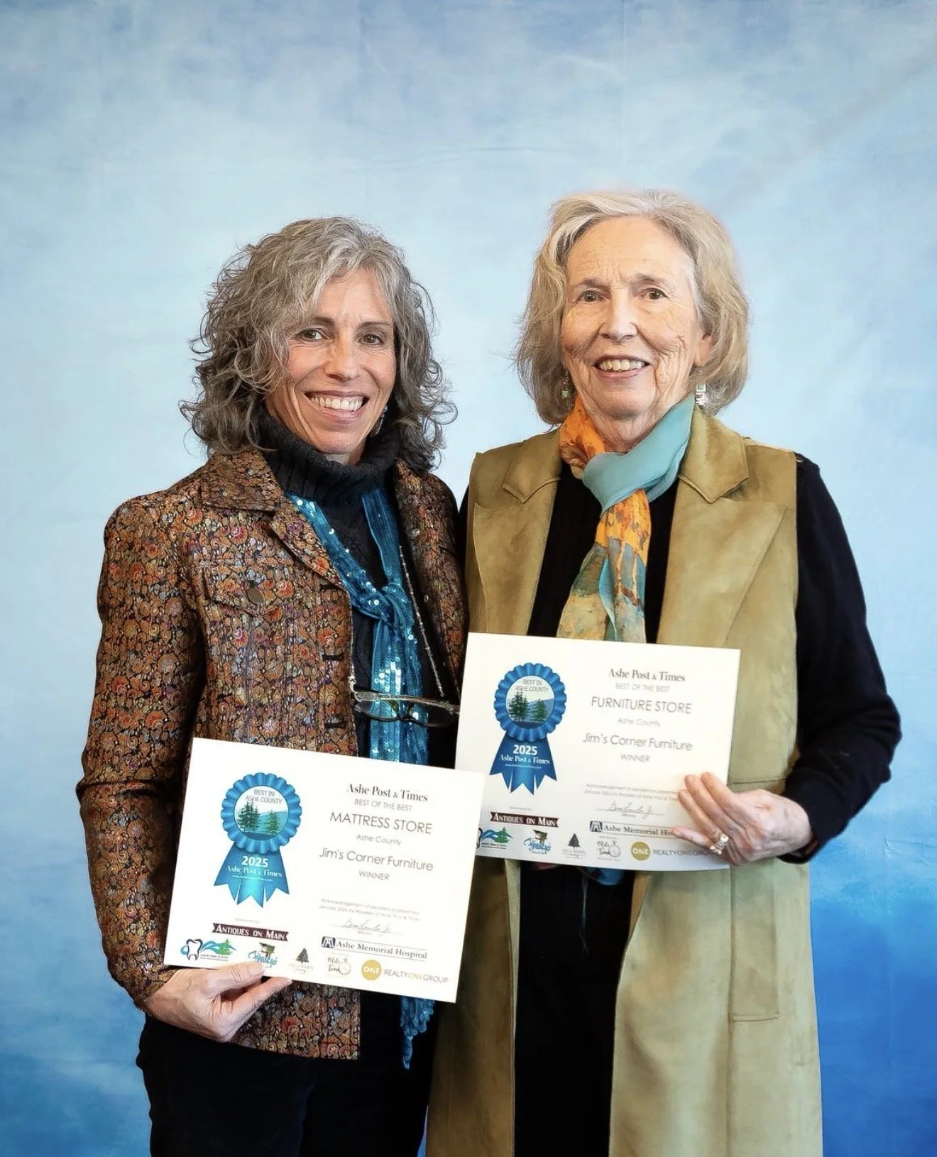 Two women smiling and holding awards for furniture store and mattress store in front of a blue backdrop.