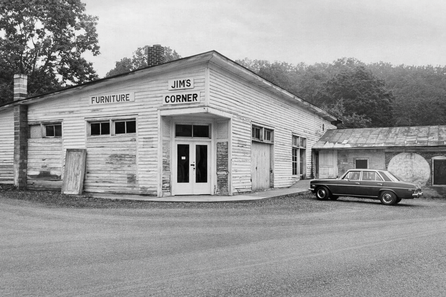 An old black and white photo of a weathered wooden building labeled "Jim's Furniture Corner" with boarded windows and a closed front door. A vintage car is parked on the street in front of the building.