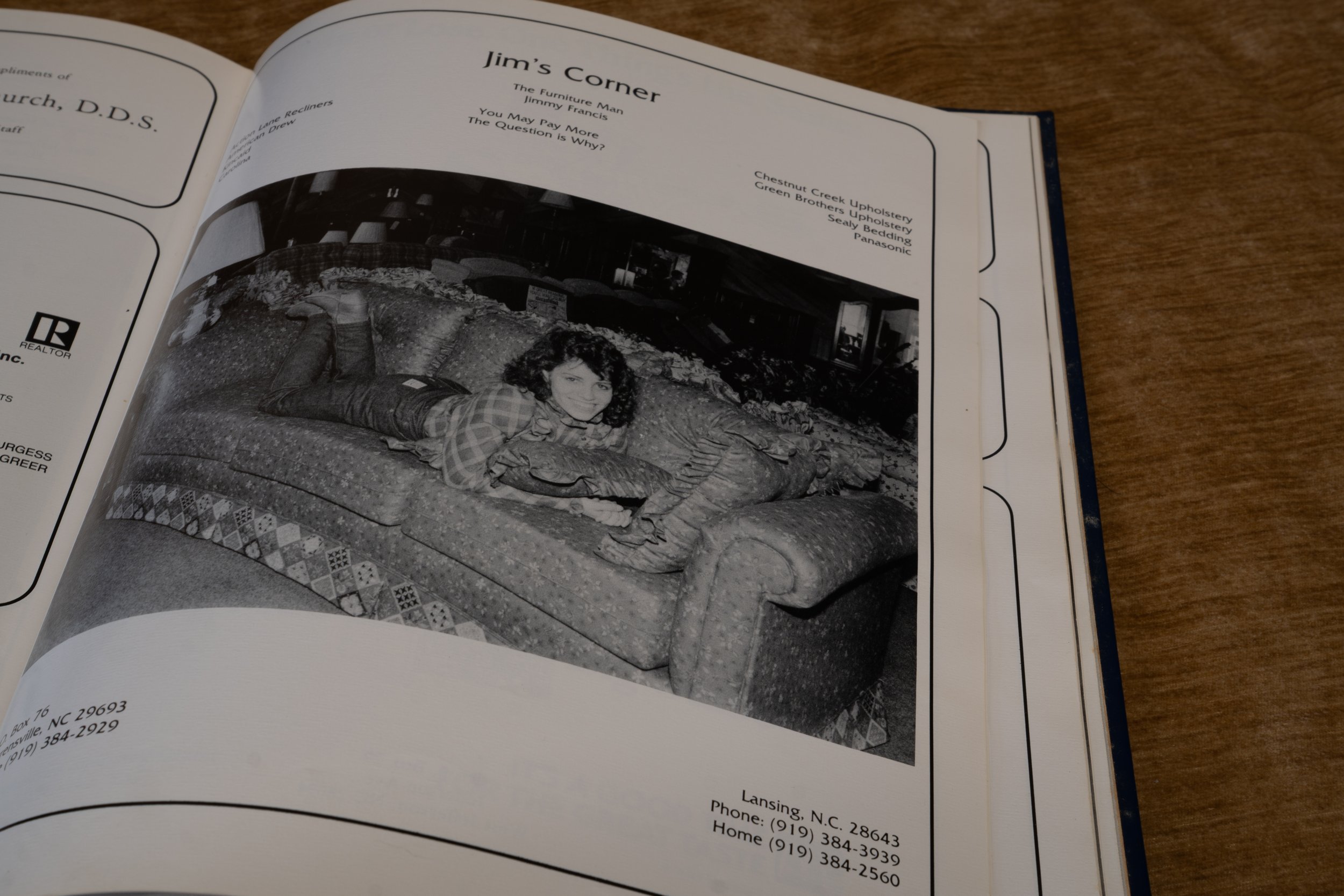 A woman lying on a patterned couch smiling at the camera in a living room with visible lamps and furniture in the background.