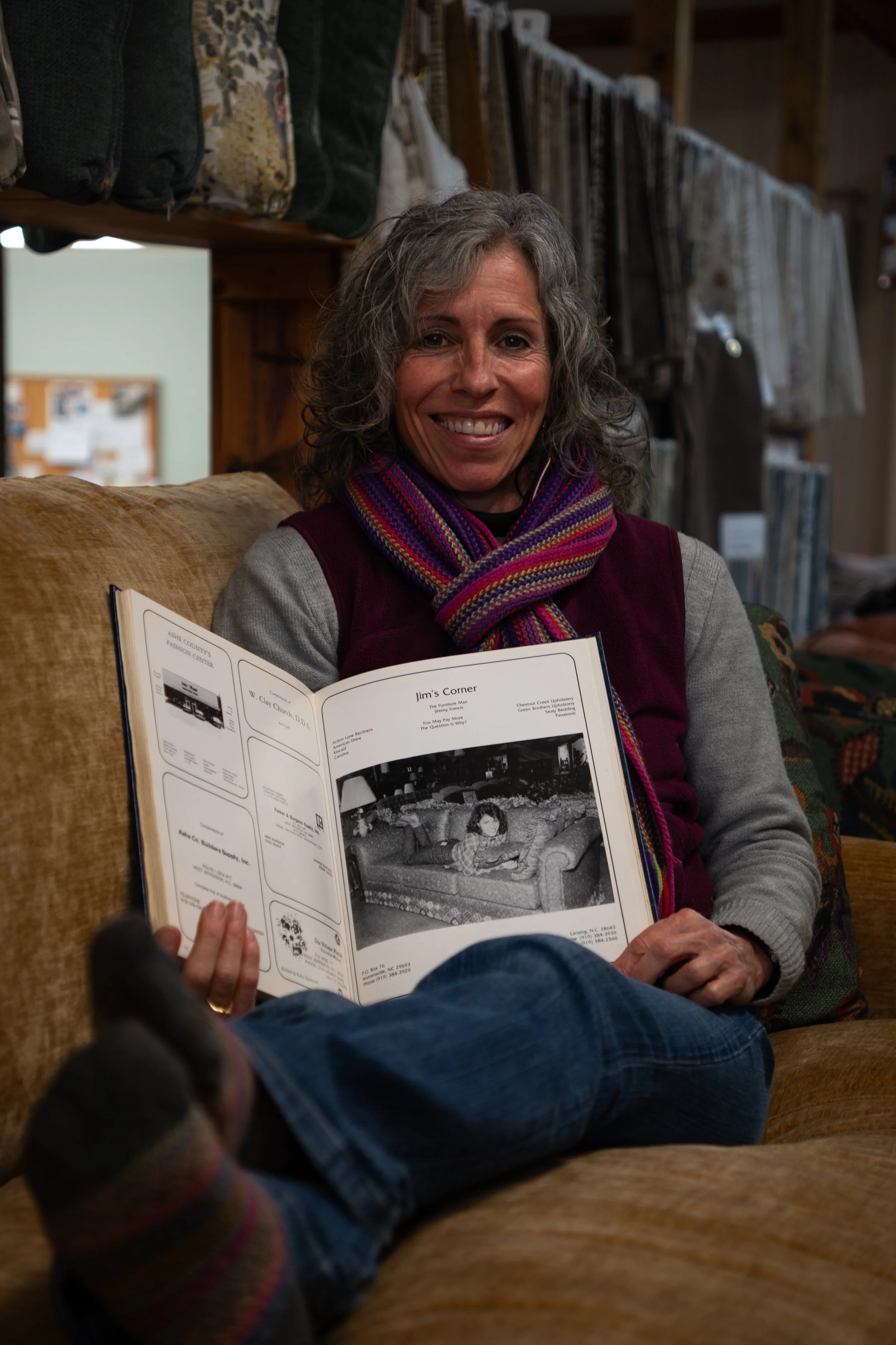 A smiling woman with curly gray hair sitting on a beige couch, holding a photo album open, with a colorful scarf around her neck. Behind her, a wooden shelf displays bags and textiles.